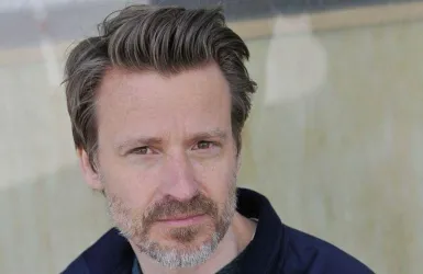 Ein Mann mit kurzen, grauen Haaren und einem Bart schaut in die Kamera. Er trägt ein dunkelblaues Oberteil und hat einen nachdenklichen Ausdruck.