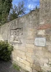 Eine steinerne Wand mit einem Relief und einer erklärenden Tafel. Im Hintergrund sind Bäume und ein blauer Himmel zu sehen.