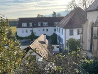 Ein charmantes altes Gebäude mit weißen Wänden und einem roten Dach in ländlicher Umgebung. Im Hintergrund sind Bäume und ein sanfter Hügel sichtbar.