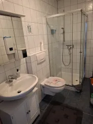 Ein einfaches Badezimmer mit einer Dusche, einem Waschbecken und einer Toilette. Die Wände sind gefliest und ein Handtuchhalter ist sichtbar.