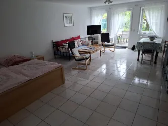 Ein helles, geräumiges Zimmer mit Fliesenboden und großen Fenstern. Es gibt eine Sitzecke, ein Bett und einen Fernseher.