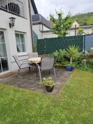 Ein gemütlicher Gartenbereich mit einem Tisch und Stühlen auf einer Terrasse. Umgeben von Pflanzen und einer grünen Wiese.