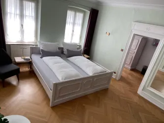 Ein helles Schlafzimmer mit einem großen, bequemen Bett und eleganten Möbeln. Neben dem Bett gibt es einen Stuhl und einen großen Spiegel.