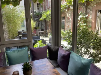 Ein stilvoller Raum mit großen Fenstern und bunten Kissen. Im Hintergrund ist ein gepflegter Garten mit Pflanzen zu sehen.