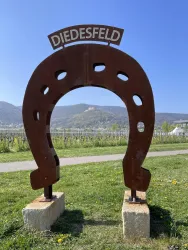 Ein großes, rostiges Hufeisen mit der Aufschrift "Diedesfeld" steht auf einem grünen Platz. Im Hintergrund sind sanfte Hügel und blauer Himmel zu sehen.