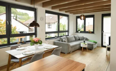 Ein modernes und helles Wohnzimmer mit großen Fenstern und einer gemütlichen Couch. Der Essbereich ist elegant gedeckt und bietet eine angenehme Atmosphäre.