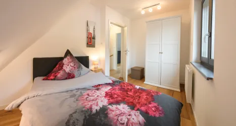 Ein gemütliches Schlafzimmer mit einem Bett, das mit floralem Bettzeug dekoriert ist. Es gibt einen kleinen Kleiderschrank und eine helle Atmosphäre durch das Tageslicht.