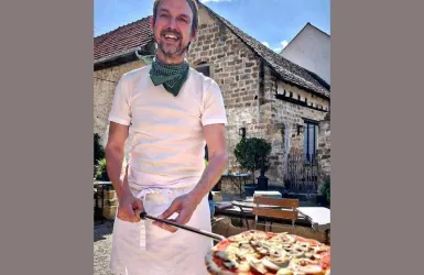 Ein jovialer Mann in Kochkleidung steht draußen und hält eine Pizzaschaufel mit einer frisch zubereiteten Pizza. Im Hintergrund ist ein charmantes Gebäude und ein gemütlicher Außenbereich zu sehen.