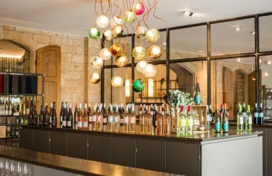 Ein moderner Weinsalon mit einer Vielzahl von Weinflaschen auf einer dunklen Theke. Über der Theke hängen stilvolle, bunte Lampen.