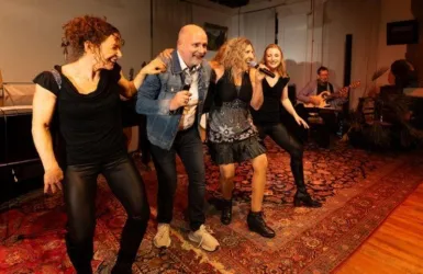 Eine fröhliche Bühnenaufführung mit vier Personen, die singen und tanzen. Im Hintergrund spielt eine Band Musik.
