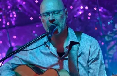 Ein Musiker spielt Gitarre und singt in einer bunten Umgebung. Im Hintergrund sind funkelnede Lichter zu sehen.