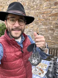 Ein Mann mit Brille und einem Hut hält ein Weinglas in der Hand. Im Hintergrund gibt es eine Tischdecke mit verschiedenen Leckereien und Gläsern.