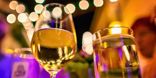 Ein elegantes Glas mit Weißwein und ein weiteres Glas mit einem kohlensäurehaltigen Getränk stehen auf einem Tisch. Im Hintergrund funkeln Lichter und schaffen eine festliche Atmosphäre.