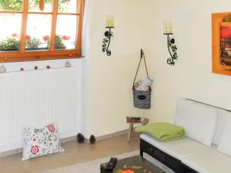 Ein gemütliches Wohnzimmer mit einem weißen Sofa und einem bunten Teppich. Die Fenster sind geöffnet und zeigen Blumenkästen draußen.