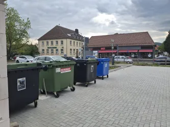 Eine Reihe von Mülltonnen steht auf einem gepflasterten Platz. Im Hintergrund sind Fahrzeuge und ein Gebäude zu sehen.