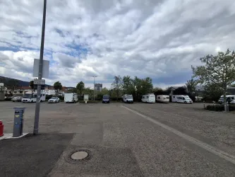 Ein Parkplatz mit mehreren Wohnmobilen und einer abwechslungsreichen Wolkenlandschaft. Im Hintergrund sind einige Bäume und Gebäude zu sehen.