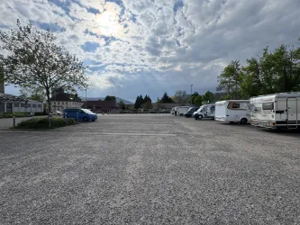 Ein großer, leerer Parkplatz mit mehreren Campingfahrzeugen und Bäumen. Der Himmel ist bewölkt, und die Umgebung wirkt ruhig.