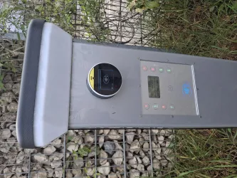 Ein Parkscheinautomat mit verschiedenen Tasten und einem digitalen Display. Er steht umgeben von Gras und Kies.