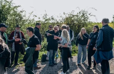 Eine Gruppe von Menschen steht im Freien und unterhält sich. Die Umgebung ist grün und hell erleuchtet von der Sonne.
