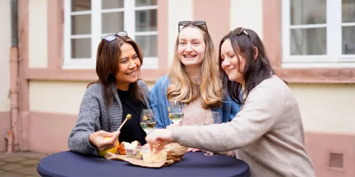 Drei Frauen sitzen an einem Tisch und genießen Snacks und Getränke. Sie lächeln und scheinen eine gute Zeit miteinander zu haben.