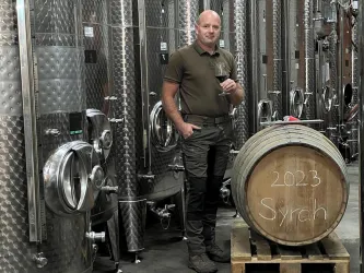 Ein Mann steht in einem Weinkeller und hält ein Glas Wein in der Hand. Neben ihm liegt ein Holzfass mit der Aufschrift "2023 Syrah".