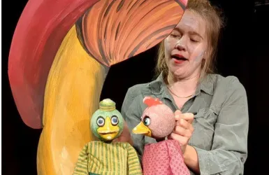 Eine Frau führt Puppen vor einem großen, bunten Hintergrund auf. Die Puppen sind ein grüner Vogel und eine rosa Henne.