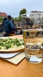 Ein Glas Wasser steht neben einer Pizza mit frischem Gemüse auf einem Holztisch. Im Hintergrund sind Menschen zu sehen, die sich im Freien entspannen.