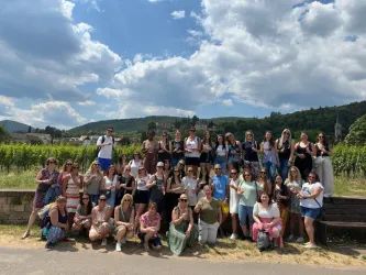 Eine große Gruppe von Menschen steht glücklich in einem Weinberg. Im Hintergrund sind sanfte Hügel und ein blauer Himmel mit Wolken zu sehen.