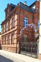 Ein elegantes, rotes Gebäude mit schönen Fenstern und einem schmiedeeisernen Tor. Vor dem Gebäude blühen lila Blumen, die eine freundliche Atmosphäre schaffen.