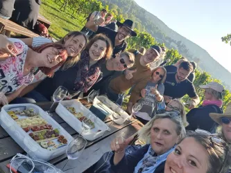 Eine Gruppe von Freunden posiert fröhlich für ein Foto in einem Weinberg. Auf dem Tisch liegen Köstlichkeiten und Gläser mit Wein.
