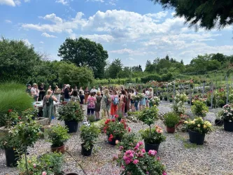 Eine Gruppe von Menschen steht in einem blühenden Garten mit vielen Pflanzen und Blumen. Im Hintergrund sind Bäume und ein blauer Himmel sichtbar.