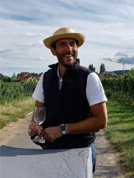 Ein Mann mit einem Strohhut und einem Glas in der Hand steht lächelnd in einem Weinberg. Im Hintergrund sind Weinreben und ein blauer Himmel zu sehen.