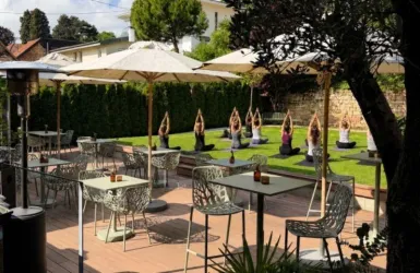 Eine ruhige Außenfläche mit Tischen und Stühlen. Im Hintergrund praktizieren mehrere Personen Yoga unter Sonnenschirmen.