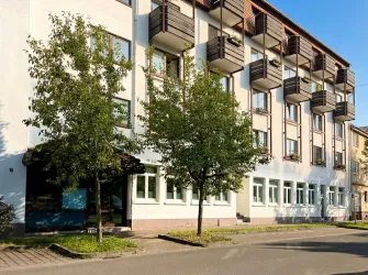 Ein modernes Gebäude mit mehreren Balkonen und großen Fenstern. Vor dem Gebäude stehen zwei kleine Bäume.