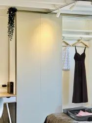 Ein modernes Schlafzimmer mit einem Kleiderschrank und einem Schreibplatz. Ein schwarzes Kleid hängt an einem Kleiderbügel, während eine Pflanze dekorativ an der Wand hängt.