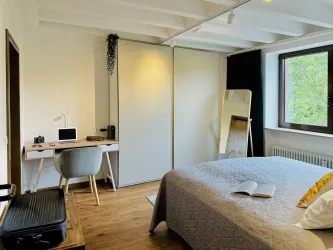 Ein helles Schlafzimmer mit einem großen Bett und einem Schreibtisch. In der Ecke steht ein großer Spiegel und es gibt einen Blick auf die Natur durch das Fenster.