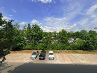 Ein Parkplatz mit mehreren Autos vor einer grünen Waldlandschaft. Der Himmel ist blau mit einigen weißen Wolken.