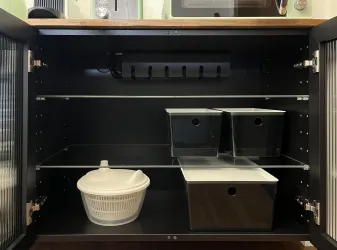 Ein leerer Schrank mit zwei Regalen. Darin befinden sich ein weißer Sieb und zwei schwarze Aufbewahrungsboxen.