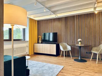 Ein modernes Wohnzimmer mit Holzverkleidung und einem großen Fernseher. Es gibt einen runden Tisch und zwei gemütliche Stühle, umgeben von hellen Farben und natürlichem Licht.