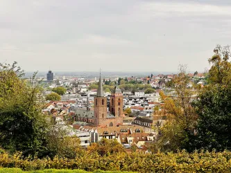 Eine Stadtansicht mit einem historischen Kirchengebäude im Vordergrund. Der Himmel ist bewölkt und die Umgebung ist grün und hügelig.