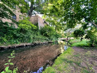 Ein ruhiger Bach fließt neben üppigem Grün und hohen Bäumen. Im Hintergrund sind alte Backsteinmauern sichtbar.