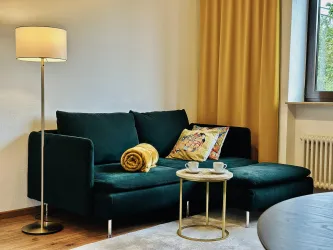 Ein gemütliches Wohnzimmer mit einem grünen Sofa und einer goldenen Decke. Neben dem Sofa steht ein Beistelltisch mit zwei Tassen und einer Stehlampe.