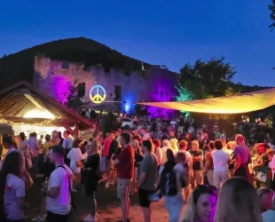 Eine lebhafte Veranstaltung bei Nacht mit vielen Menschen und bunten Lichtern. Im Hintergrund ist eine historische Burg mit einem Friedenszeichen beleuchtet zu sehen.