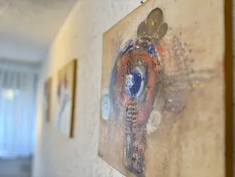 Ein Kunstwerk an einer Wand mit bunten Farben und abstrakten Formen. Im Hintergrund sind weitere Bilder und ein Fenster zu sehen.