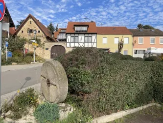 Ein traditionelles Gebäude aus Fachwerk steht neben modernen Häusern in bunten Farben. Im Vordergrund liegt ein großer Stein, umgeben von grünen Sträuchern.