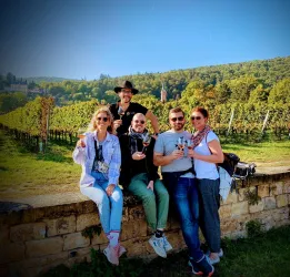 Eine Gruppe von Freunden steht vor einer Weinberglandschaft und hält Gläser in der Hand. Im Hintergrund sind grüne Hügel und Bäume sichtbar.