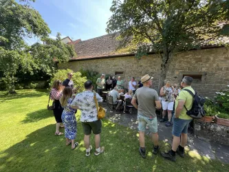 Eine Gruppe von Menschen steht im Freien in einem Garten. Im Hintergrund ist eine steinerne Wand und Bäume zu sehen.