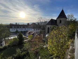 Eine malerische Aussicht auf eine Kirche und umliegende Gebäude im Sonnenlicht. Im Vordergrund sind grüne Pflanzen und Bäume zu sehen.