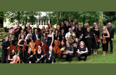 Eine große Gruppe von Orchestermusikern steht im Freien unter Bäumen. Die meisten tragen schwarze Kleidung und lächeln in die Kamera.