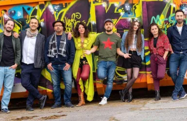Eine Gruppe von acht Personen steht vor einer bunten Graffiti-Wand. Sie lächeln und zeigen unterschiedliche Mode-Stile.